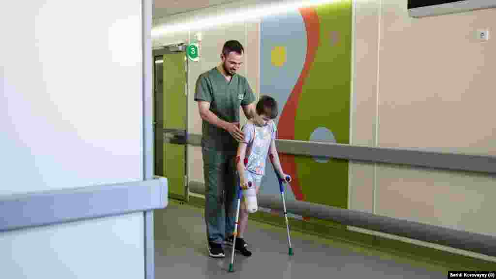 Saško, 11-godišnji dječak koji je preživio napad granatiranjem, hoda pomoću štaka dok mu pomaže član osoblja bolnice Ohmadit u martu 2024. U januaru 2024. Saško je sa sestričnom Alinom išao do lokalne trgovine u regiji Harkiv kada je u blizini eksplodirala granata. Eksplozija je dječaku otrgnula nogu i teško ranila Alinu, koja je umrla nakon 10 dana na intenzivnoj njezi.  