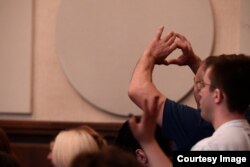 დამსწრე შორიდან ესალმება ბრალდებულებს და ხელით აჩვენებს გულის ფორმას.