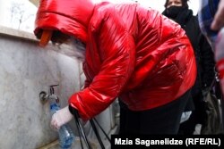 ქალი დამცავი ფარით, ნიღბით და ხელთათმანით ნაკურთხ წყალს იმარაგებს.