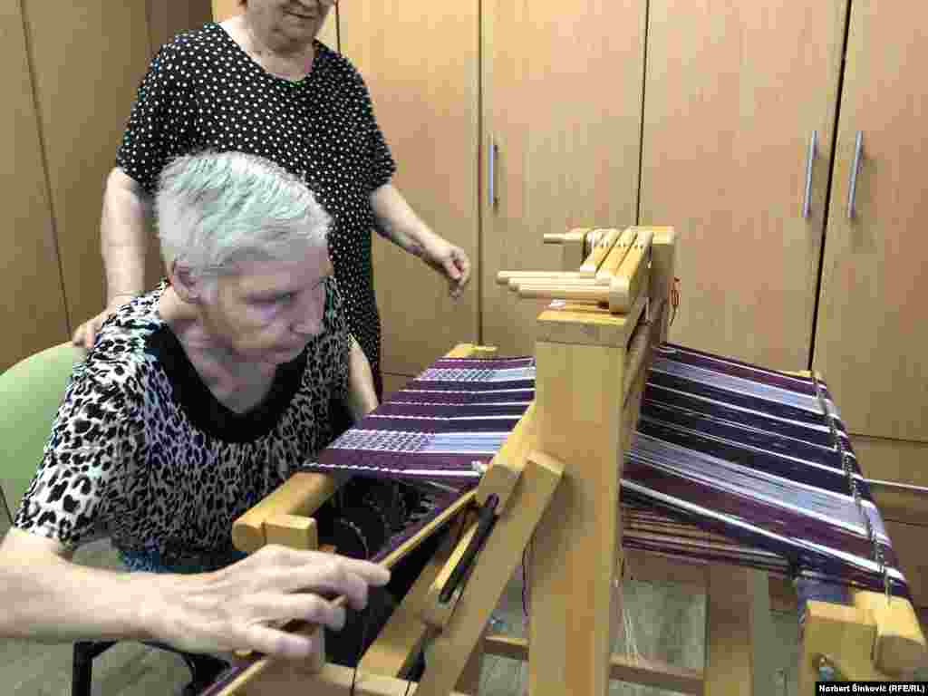 Među njima je i Maša, koja uz pomoć Ljiljane Živković uči kako ručno tkati. Ljubičasti pamuk i svileni konac očitavaju složenu formu na tekstilu - pravi se pončo za leto. „Ovde stvarno nije ništa teško“, kaže sagovornica RSE iz Udruženja za pomoć mentalno nedovoljno razvijenim osobama.