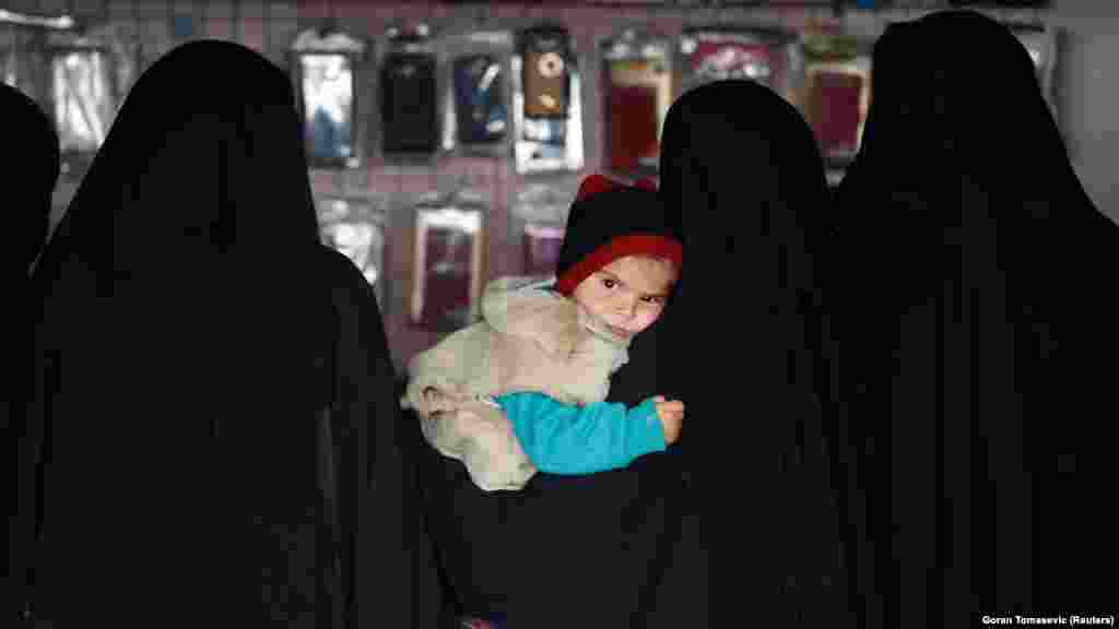 Žena nosi dijete u trgovini u kampu Al-Hol, 8. siječnja 2020. Većina žena s kojima je Reuters pokušao razgovarati, odbila je govoriti. Ona koja je pristala pričati nije kazala svoje ime. Govorila je na lošem engleskom i rekla da je porijeklom iz Hong Konga. Istakla je da je došla na Bliski istok kako bi se pridružila "Islamskoj državi". "Imam jedno dijete, a muž mi je umro u Baguzu", rekla je. Grad Baguz, posljednja enklava "Islamske države" na istoku Sirije, pao je u proljeće 2019. godine od kurdskih i američkih snaga. Žena je rekla da je u kontaktu sa svojom obitelji u Hong Kongu, ali da se ne želi vratiti. "Znam da je ovdje situacija vrlo teška. Ovo nije dom, to je samo šator... Ali svi živimo prema (Allahovim) željama. Tako da - ako je Bog tako htio, onda je sve dobro."